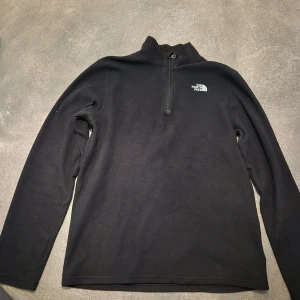 Svart fleece-tröja från The North Face - Svart fleece-tröja från The North Face med halv dragkedja och hög krage. Perfekt för kyliga dagar, enkel och stilren design med logga på bröstet.