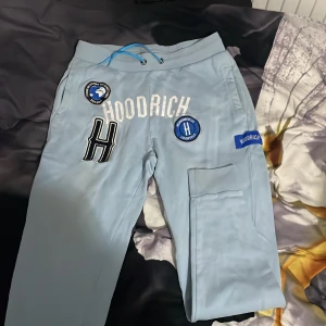 Ljusblå Hoodrich mjukisbyxor med patchar - Säljer ett par ljusblå mjukisbyxor från Hoodrich med flera coola patchar och broderade detaljer framtill. Byxorna har snörning i midjan och en praktisk cargoficka på sidan. Perfekta för en avslappnad streetwear-look.