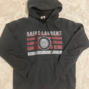 Saint Laurent Hoodie - Snygg svart Saint Laurent-hoodie med vintage-inspirerat tryck fram. Skön passform och exklusiv känsla — klassisk men ändå street. Använd få gånger, i mycket bra skick den är strlk M men känns mer som S