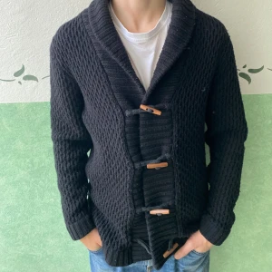 Svart kofta från These Glory Days - Svart grovstickad kofta från These Glory Days i storlek medium. Koftan har bred krage och snygga träknappar framtill som ger en cool touch. Långärmad och med ribbade muddar vid ärmslut och nederkant. Perfekt för dig som gillar stilren och enkel design.