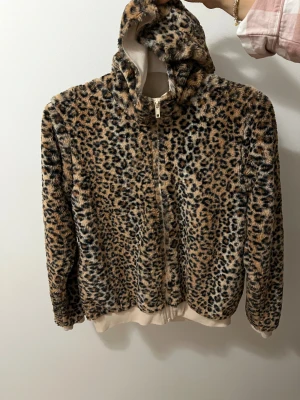 Leopardmönstrad teddyjacka med huva - Säljer en fluffig teddyjacka med leopardmönster och dragkedja framtill. Jackan har huva och mjuka muddar i beige vid ärmslut och nederkant. Perfekt för dig som vill sticka ut med en cool och djurinspirerad look.