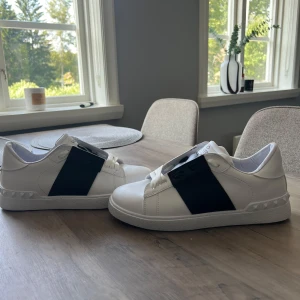Valentino Garavani Open sneakers vit/svart k0pia - Snygga Valentino Garavani Open sneakers i vitt skinn med bred svart rem över vristen. Klassisk rund tå, vita snören och coola nitar längs sulan. Perfekt för dig som vill ha en stilren men ändå unik look. Sulan har mönstrad struktur och logga undertill. Otroligt bra skick aldrig använda 