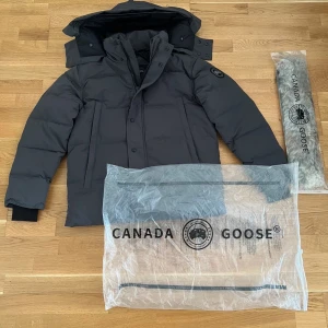 Canada Goose Wyndham  - Säljer en svart dunjacka från Canada Goose med avtagbar huva och pälsdetalj. Jackan har snygga svarta knappar, dragkedja och logotyp på ärmen. Perfekt för kalla vinterdagar och har en stilren, puffad look.