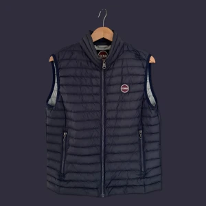 Mörkblå dunväst från Colmar - Colmar väst i mörkblå färg. Västen har en dragkedja fram och två fickor med dragkedja. Colmar-logotypen finns på vänster bröst. Mycket gott skick, västen är väl omhändertagen. Storlek 48 (Medium). Nypris ca 3000kr.