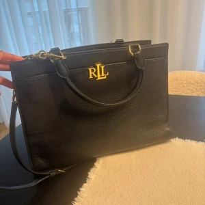 Svart handväska från Ralph Lauren - Stilren svart handväska i skinn från Ralph Lauren med guldfärgade detaljer och logotyp framtill. Väskan har både handtag och axelrem, samt flera fack och dragkedja upptill. Perfekt för dig som vill ha en klassisk och snygg accessoar. Köpt för 2500. Säljer för 1000 pris kan diskuteras .