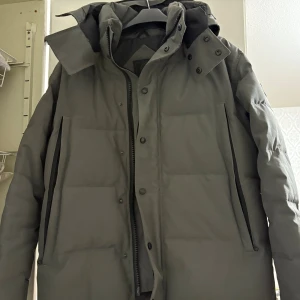 Grå dunjacka från Canada Goose M - Säljer en grå dunjacka från Canada Goose i storlek M. Jackan har huva, svarta dragkedjor på fickorna och knappar framtill. Klassisk patch på ärmen och quiltad design. Perfekt för kalla dagar med stilren look.