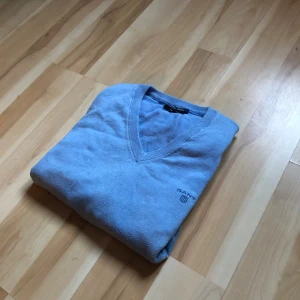 Ljusblå v-ringad tröja från GANT - Snygg ljusblå v-ringad tröja från GANT i premium bomull. Tröjan har lång ärm, ribbade muddar och diskret broderad logga på bröstet. Perfekt för dig som gillar klassisk stil med en modern touch.