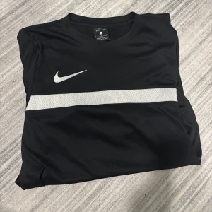 Svart Nike Dri-Fit fotbollströja S - Snygg svart fotbollströja från Nike med Dri-Fit-teknologi. Tröjan har en vit rand över bröstet och klassisk Nike-logga. Tillverkad i lätt och ventilerande polyester, perfekt för träning eller match. Kortärmad och sportig design.