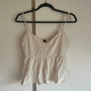 Vit randig linnetopp med v-ringning - Säljer en söt vit linnetopp med tunna axelband och v-ringning. Toppen har ett subtilt randigt mönster och en lätt utställd passform nedtill. Perfekt för varma dagar och enkel att styla med jeans eller kjol.
