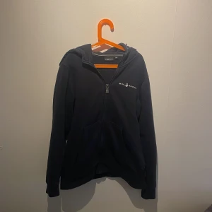 Blå hoodie med dragkedja från Sail Racing - Blå hoodie från Sail Racing med dragkedja framtill och två fickor. Tröjan har broderad logga på bröstet och en detalj på huvan. 