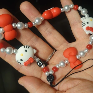 Hello Kitty, Sanrio, Telefonrem, mobilsmycke, nytt! - Supergulligt mobilsmycke med Hello Kitty-huvuden, röda rosetter och pärlliknande kulor i silver och rött. Armbandet har detaljer i mjuk plast och elastisk tråd, perfekt för dig som älskar kawaii-stil och vill sticka ut med något lekfullt och färgglatt. 15 cm långt.