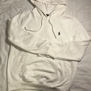 Vit hoodie från Polo Ralph Lauren - Vit hoodie från Polo Ralph Lauren med klassisk broderad logga på bröstet. Tröjan har huva med snörning, stor magficka och ribbade muddar. Mjuk bomullskvalitet och stilren design som funkar till allt. Perfekt för dig som gillar clean och enkel stil.