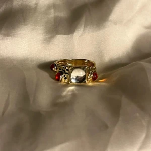 Guldfärgad ring med röda stenar - Unik guldfärgad ring med en stor fyrkantig klar sten i mitten och tre runda röda stenar på sidorna. Ringen har detaljerade utskärningar i metallen som ger en cool och lyxig vibe. Perfekt för dig som gillar statement-smycken och vill sticka ut.