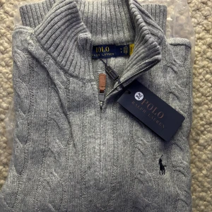 Grå stickad polotröja Polo Ralph Lauren - Kabelstickad grå polotröja från Polo Ralph Lauren med half zip och ribbade muddar. Klassisk design med broderad logga på bröstet och hög krage. Perfekt för lager-på-lager och snygg till jeans eller chinos.