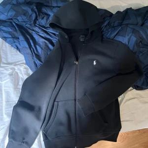 Svart hoodie från Polo Ralph Lauren - Svart hoodie från Polo Ralph Lauren med dragkedja och klassisk vit logga på bröstet. Tröjan har huva, känguruficka och är tillverkad i mjuk bomullsmix. Perfekt för en chill och stilren look. 