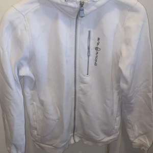 Vit Sail Racing vindjacka med huva - Snygg vit hoddie Sail Racing med huva och dragkedja framtill. Jackan har en extra bröstficka med dragkedja och logga på både bröstet och huvan. Perfekt för dig som gillar sportig stil och vill ha en jacka som funkar till många tillfällen. Obs! Som ni ser på bilden finns det en fläck som jag inte fått bort i tvätten. Förövrigt är den hel.