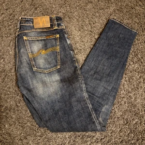 Nudie Jeans Skinny Lin blå jeans - Säljer ett par Nudie Jeans Skinny Lin i tvättad blå färg med snygga slitningar och klassiska orange sömmar. Modellen är skinny fit med smal passform och tillverkad av 98% ekologisk bomull och 2% elastan för extra stretch. Bakfickan har Nudies ikoniska brodyr.