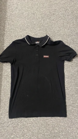 McKenzie Polo Shirt - Svart pikétröja från McKenzie med vit piping på kragen och tryckt logga på bröstet. Axlarna har ett band med McKenzie-tryck för extra streetkänsla. Tröjan har korta ärmar och knappar framtill. Materialet är mjuk bomull som är skön mot huden.