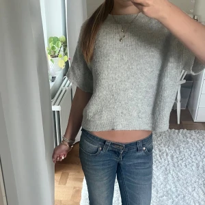 Grå croppad stickad tröja - Supermysig ljusgrå stickad tröja med ribbad struktur och croppad passform. Tröjan har korta ärmar och rund halsringning, perfekt att styla med jeans eller kjol. Materialet är mjukt och ger en chill vibe.