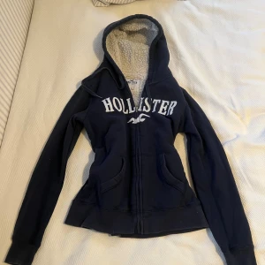 Mörkblå Hollister hoodie med teddyfoder - Mysig mörkblå hoodie från Hollister med vit logotyp framtill. Tröjan har huva med snörning, dragkedja och två fickor. Insidan är fodrad med fluffigt teddyfoder som gör den extra varm. Perfekt för kyliga dagar och chill stunder.