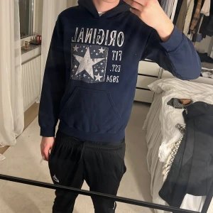 Mörkblå hoodie med stjärntryck - Snygg mörkblå hoodie från Original Fit med stort vitt stjärntryck och texten 'Original Fit Est. 2024' på bröstet. Tröjan har huva, magficka och långa ärmar. Perfekt för chill dagar och streetstyle. Modellen är 184 cm lång och väger 72 kg. 