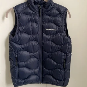 Mörkblå dunväst Peak Performance - Snygg mörkblå dunväst från Peak Performance med quiltad design och hög krage. Västen har två praktiska fickor med dragkedja och ett broderat logotyp på bröstet. Perfekt för lager-på-lager och outdoor-aktiviteter.