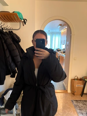 Svart pufferjacka med bälte - Säljer en svart pufferjacka med bälte i midjan och hög krage. Jackan har lång ärm och en oversized passform som ger en cool och avslappnad look. Perfekt för kalla dagar när du vill vara både varm och snygg.
