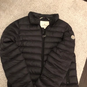 Svart Moncler dunjacka herr - Svart dunjacka från Moncler med klassisk quiltad design och dragkedja framtill. Jackan har lång ärm, ståkrage och Moncler-logga på vänster ärm. Insidan har en cool serietidningsdetalj och en röd dragkedja på innerfickan. Perfekt för kalla dagar.