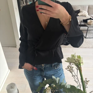 Svart omlottblus från Bik Bok - Säljer en svart omlottblus från Bik Bok i storlek M. Blusen har långa ärmar, v-ringad hals och knyt i sidan som ger en snygg siluett. Materialet är mjukt och har en lätt glans. Perfekt att styla med jeans eller kjol för en cool look.