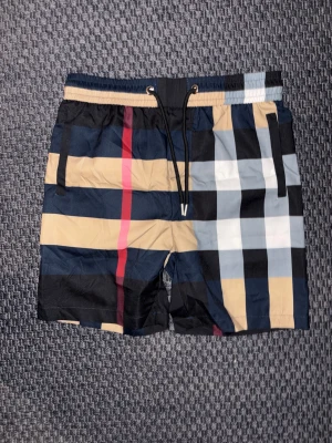 Rutig Burberry shorts med snörning storlek S inte M! - Säljer ett par snygga Burberry shorts med klassiskt rutigt mönster i beige, svart, blått, vitt och rött. Shortsen har elastisk midja med svart snörning och sidofickor. Perfekta för sommaren och ger en clean och trendig look.