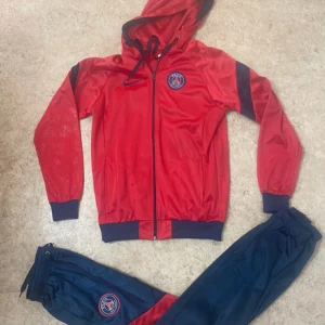 PSG röd och blå träningsset Nike - Snyggt träningsset från PSG och Nike med röd huvtröja med dragkedja och blå detaljer samt matchande blå byxor med PSG-logga. Tröjan har huva med snörning och muddar vid ärmslut och midja. Byxorna har resår och snörning i midjan. Perfekt för dig som gillar fotboll och sportig stil.