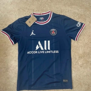 PSG marinblå fotbollströja Jordan - Snygg PSG fotbollströja i marinblått med vita och röda detaljer på krage och ärmslut. Tröjan har korta ärmar, PSG-logga på bröstet och Jordan-logga. Materialet är lätt och andas, perfekt för match eller träning. Tryck med 'Accor Live Limitless' på framsidan.