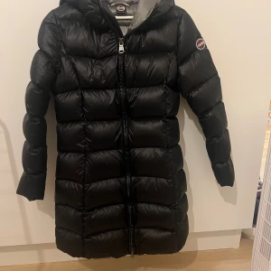 Svart lång dunjacka från Colmar - Lång svart dunjacka från Colmar med huva och quiltad design. Jackan har dragkedja framtill och logga på ärmen. Perfekt för kalla dagar och riktigt snygg siluett. Fodrad och fluffig för extra värme. Colmars storlek motsvarar S. Som ny. Använt endast ett par ggr. 