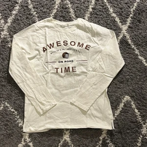 Vit långärmad tröja MNG med tryck - Vit långärmad tröja från MNG med coolt tryck på bröstet där det står 'AWESOME TIME' och en liten döskalle med hjälm. Tröjan är i mjuk bomull och har en normal passform. Perfekt för dig som gillar streetstyle och grafiska prints. Passar perfekt för 12 åringar
