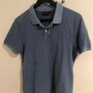 Blå pikétröja från Morris - Snygg blå pikétröja från Morris med klassisk krage och två knappar framtill. Tröjan har korta ärmar och en liten broderad logga i orange på bröstet. Tillverkad i mjuk bomull som är skön mot huden. Perfekt för en chill och stilren look.