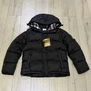 Svart Burberry jacka  - Svart dunjacka från Burberry med avtagbara ärmar, så du kan bära den som väst. Jackan har en stor huva, dragkedjor på fickorna och ett rutigt foder i grått, vitt och rött. Perfekt för kalla dagar och har en cool, puffad look.