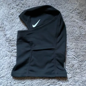 Nike Pro svart ski mask - Råka slänga förpackningen.         Svart balaclava från Nike Pro med vit swoosh-logga på sidan. Tillverkad i mjukt och stretchigt syntetmaterial som sitter tight och täcker hela huvudet och halsen. Perfekt för träning utomhus när det är kallt. Snygg, sportig look med diskret Nike Pro-märke framtill.