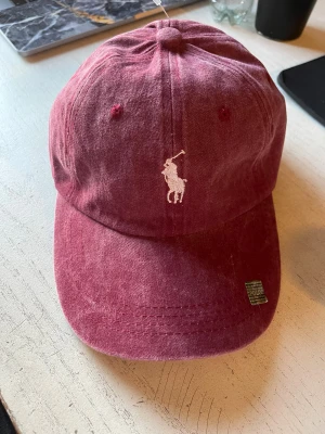 Vinröd keps från Polo Ralph Lauren - Snygg vinröd keps från Polo Ralph Lauren med klassisk broderad logga framtill och 'POLO' broderat baktill. Kepsen är tillverkad i 100% bomull och har justerbart spänne bak för perfekt passform. Perfekt accessoar för en chill och sportig look.