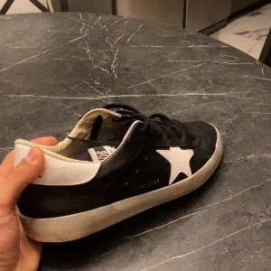 Svarta Golden Goose sneakers med stjärna - Säljer ett par svarta Golden Goose sneakers med vit stjärndetalj på sidan och vit häl. Skorna har snörning och en klassisk platt sula. Materialet är en mix av skinn och mocka, vilket ger en snygg kontrast. Perfekta för dig som gillar streetstyle och vill sticka ut.