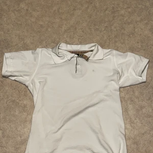 Vit pikétröja Burberry London L - Vit pikétröja från Burberry London i storlek L. Passar även M. Klassisk krage med knappar och ikoniskt rutigt tyg på insidan av kragen. Tröjan har korta ärmar och en stilren look som passar dig som gillar premium streetwear. Materialet är mjuk bomull. Använd 2 gånger