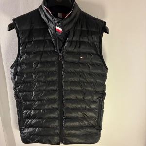 Tommy Hilfiger – Svart dunväst (stl S, stor i modellen) - Säljer en svart dunväst från Tommy Hilfiger i storlek S. Den är lite stor i modellen och passar därför bäst för någon som vanligtvis använder storlek M eller har en bredare kroppstyp. Lätt och smidig väst med dragkedja framtill och två fickor med dragkedjor, samt diskreta Hilfiger-detaljer vid bröstet och i kragen. Västen är använd men i bra skick utan skador. Nypris är 2 049 kr.