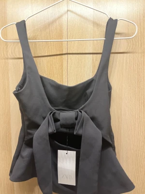 Svart topp med rosett från Zara XS - Supersnygg svart topp från Zara i storlek XS. helt oanvänd med lapp kvar. Modellen har breda axelband, figurnära passform och en stor rosett på baksidan.  Toppen är lätt utsvängd nertill och Perfekt att styla med jeans eller kjol.
