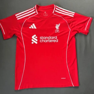 Liverpool röd fotbollströja Adidas - Röd Liverpool fotbollströja från Adidas med vita detaljer och klubbmärke på bröstet. Tröjan har klassiska vita ränder på axlarna, rund hals och tryckt sponsorlogga framtill. Tillverkad i lätt och ventilerande material.