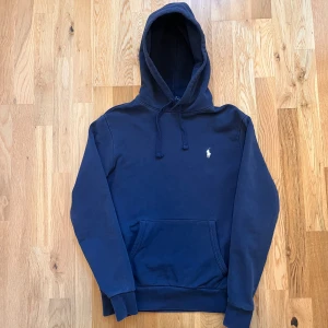 Marinblå hoodie från Polo Ralph Lauren - Säljer en stilren marinblå hoodie från Polo Ralph Lauren med klassisk broderad logga på bröstet. Tröjan har huva med snörning, känguruficka och är tillverkad i mjuk bomull. Perfekt för en chill och avslappnad look.