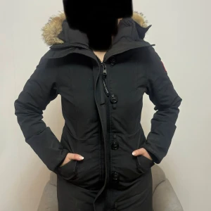 Svart parkas från Canada Goose - Svart parkas från Canada Goose med päls på huvan och klassisk logga på ärmen. Jackan har långa ärmar, två stora fickor framtill och dragkedja med knappslå. Perfekt för kalla vinterdagar och har en snygg, stilren look. Såklart äkta.