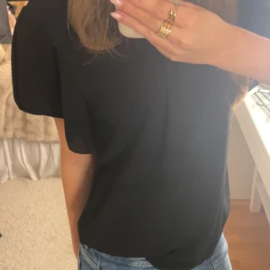 Svart blus med vida ärmar - Säljer en svart blus med rundad hals och snygga, vida korta ärmar. Blusen har en luftig passform och är tillverkad i ett lätt, flowy material som ger en skön känsla. Perfekt att styla med jeans eller kjol för en clean look.