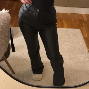 Svarta raka byxor i skinnimitation - Snygga svarta byxor i skinnimitation med raka ben och hög midja. Byxorna har klassiska fickor fram och bälteshällor, vilket ger en clean och trendig look. Perfekta för dig som vill sticka ut med en edgy stil.