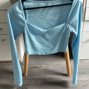 Ljusblå ribbad långärmad topp - Snygg ljusblå långärmad topp med ribbad struktur och unik skärning vid halsen. Toppen är croppad och har en smickrande passform som passar perfekt till jeans eller kjol. Perfekt för dig som vill ha en trendig och stilren look.