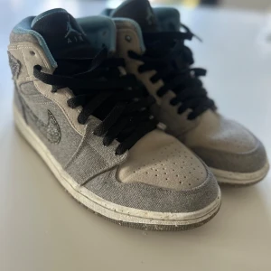 Nike Air Jordan 1 High Grå/Beige - Säljer ett par Nike Air Jordan 1 High i grått och beige med svart snörning och ljusblå insida. Ovandelen är i textil och mocka, med en unik Swoosh i mönstrad stil. Sulan är i gummi med färgade prickar för en cool look. Perfekta för dig som gillar streetwear och sneakers.
