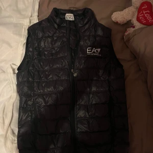 Svart dunväst från Emporio Armani EA7 - Snygg svart dunväst från Emporio Armani EA7 med quiltad design och logotyp på bröstet. Västen har dragkedja framtill och står upp lite vid kragen. Perfekt att slänga över en hoodie eller t-shirt för en cool look. passar tyvärr inte mig längre.